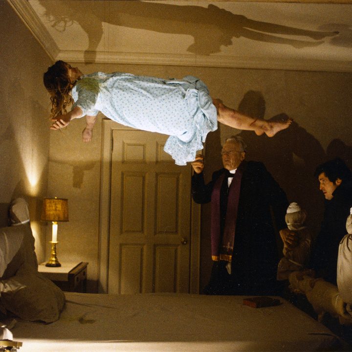 A girl levitates above her bed in front of two priests.