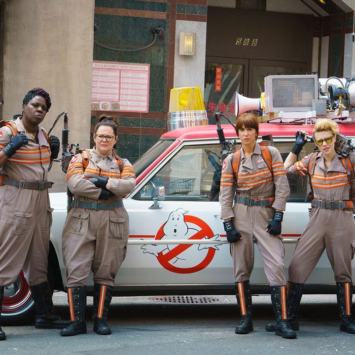 Four female ghostbusters stand in front of a car.