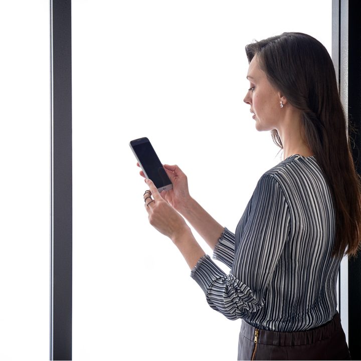 A woman standing in front of a window, looking at the smartphone in her hand.
