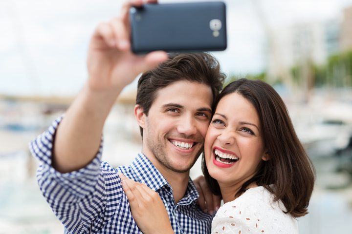young couple taking a self portrait laughing as they pose close together for the camera on their smartphone outdoors in summer sunshine