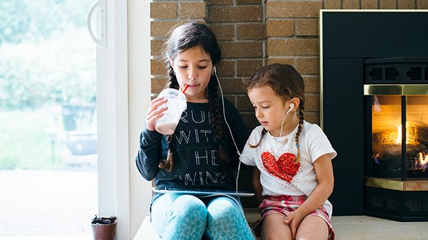 Two girls (one older, one younger) sit side by side in front of a fire, sharing a pair of wired earphones while the older girl holds a tablet and they watch the screen.