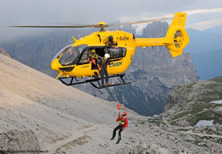 A man hangs on a cable from a yellow helicopter, with mountains in the background.