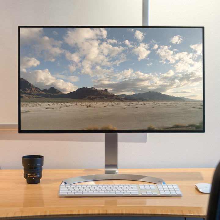 A large computer monitor and a keyboard sitting on a wooden table in front of a white wall.