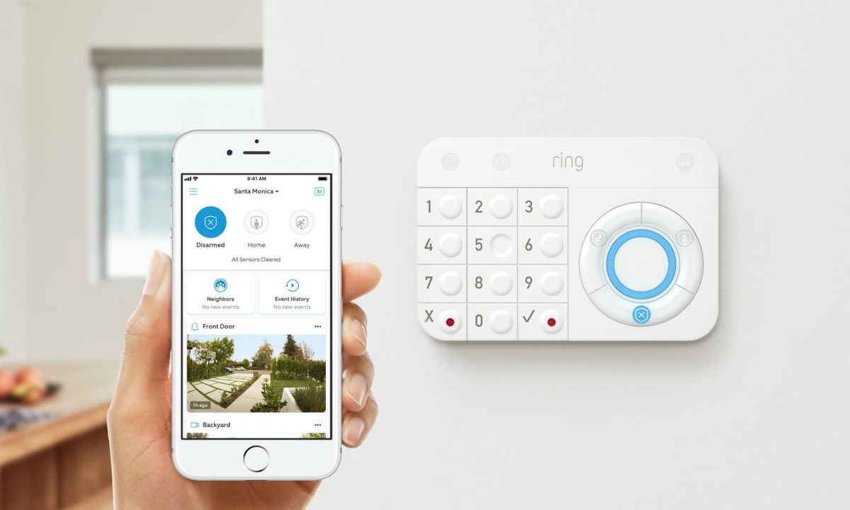 The Ring alarm on a wall with a hand holding a phone with the home app in front of it.