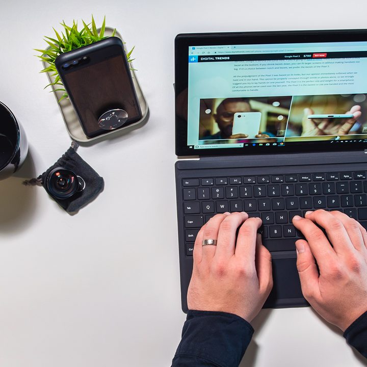 A pair of hands typing on a tablet keyboard, on a white table that also features a phone, an empty coffee cup, a camera lens, and a green plant.