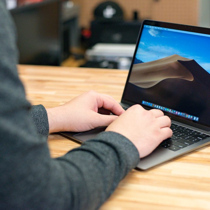 A person's hands resting on a 2018 Macbook Air as it sits on a wooden table.