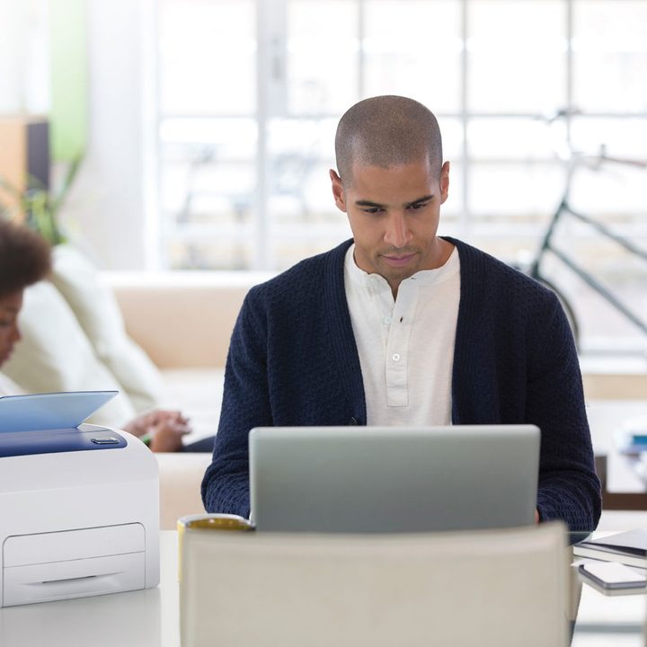 A person sitting at a table with a laptop and printer on it. Another person is sitting in the background.