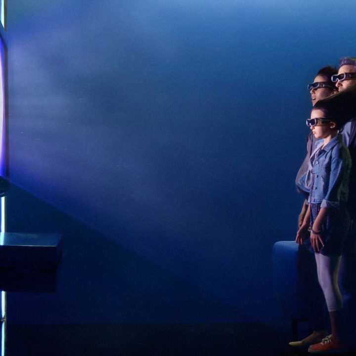 Three individuals stand in front of a large 3D TV wearing 3D glasses and having their hair blow back by the realism of the image.