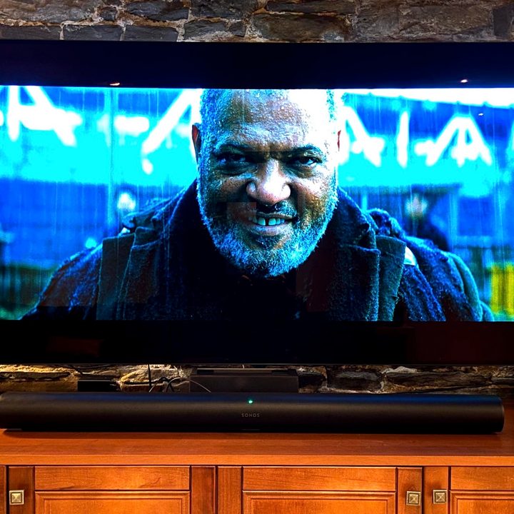 Laurence Fishburne grins into the camera displayed on a TV above a Sonos Arc Dolby Atmos soundbar.