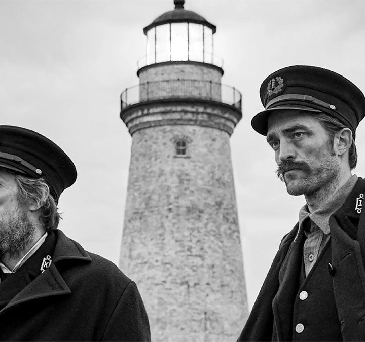 Willem Dafoe and Robert Pattinson in character with a lighthouse in the background in The Lighthouse.