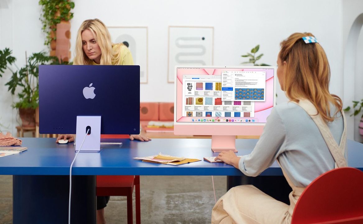 Two people use iMacs on a desk in an office.