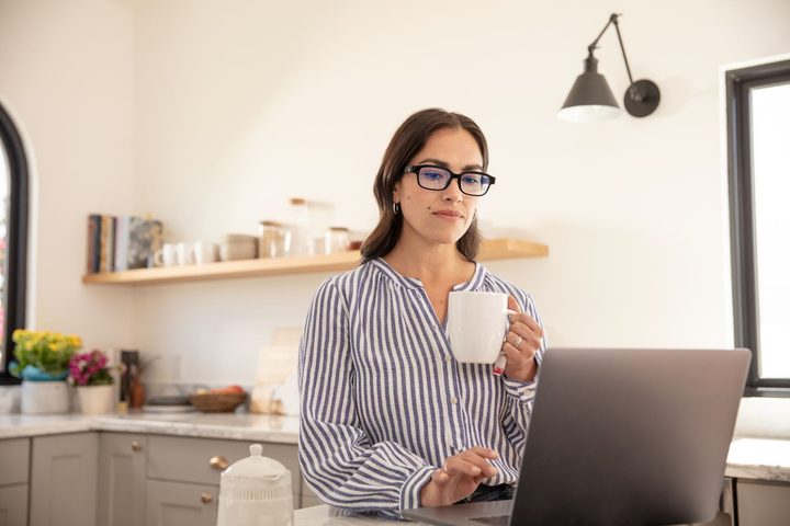 A woman wearing echo frames, sitting in front of her computer, drinking coffee.