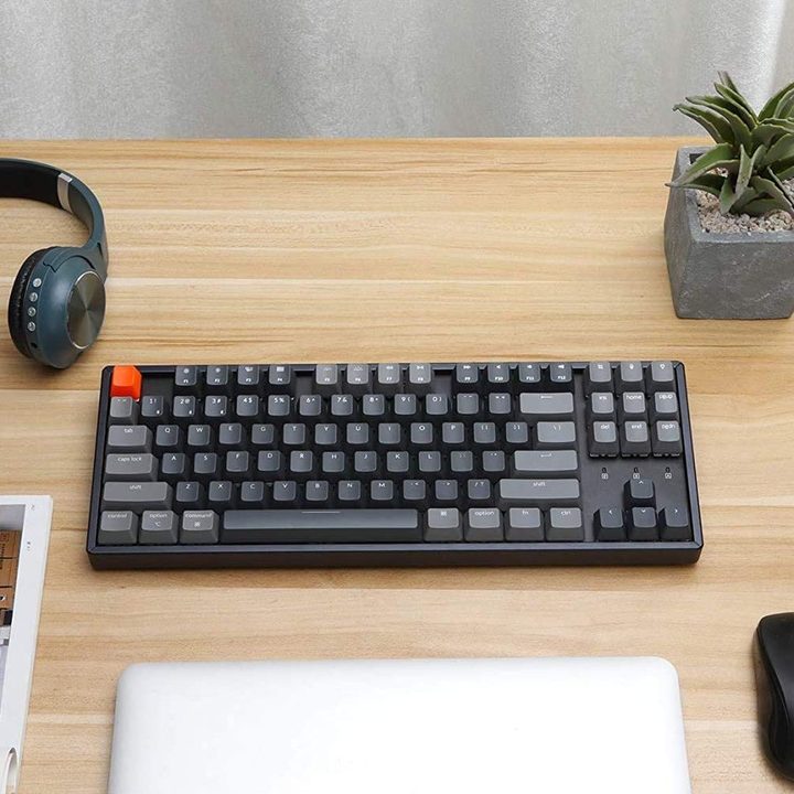 The Keychron K8 keyboard on a wooden table with a mouse, computer, and headphones.