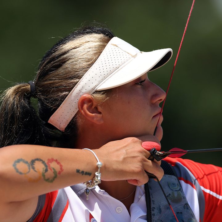 A female archer sporting a tattoo of the Olympic Rings pulls back the string of a bow with a nocked arrow.