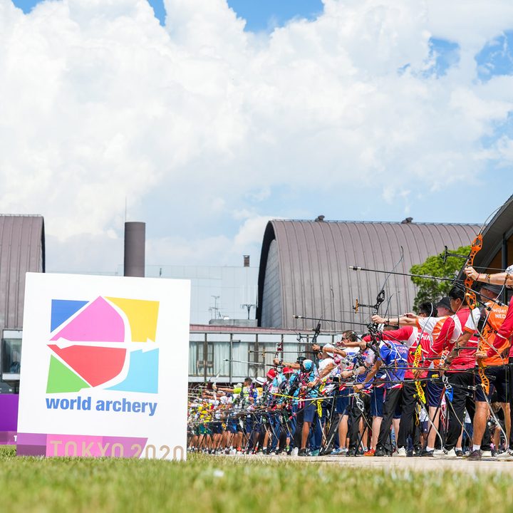 A group of archers with bows drawn aim over a sign for World Archery Tokyo 2020.