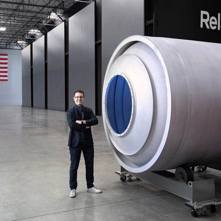 Relativity Space CEO, Tim Ellis standing next to a rocket component. An American flag is in the background.