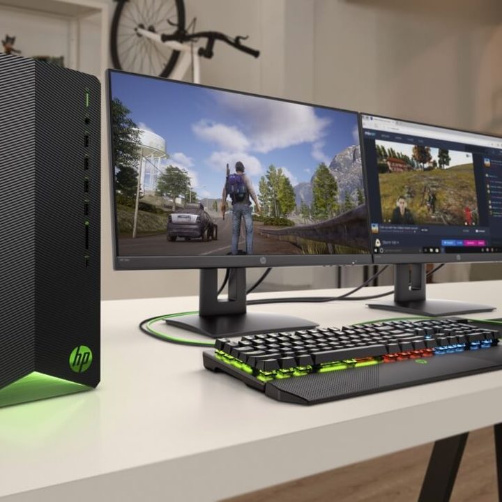 HP Pavilion Gaming Desktop on a desk with two monitors nearby displaying games and a keyboard and headset to the side.