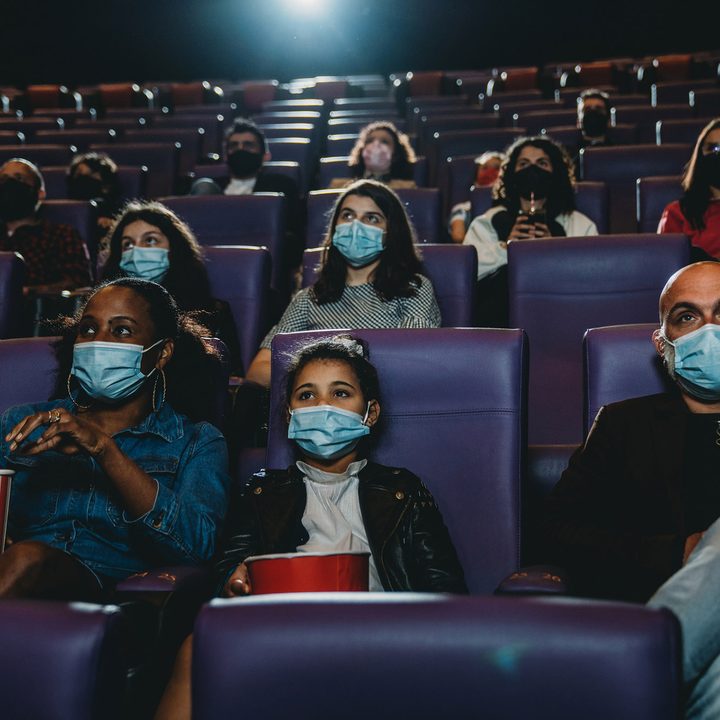 People at the movie theater watching a movie. They are wearing protective face masks. Focus on a family with one child.