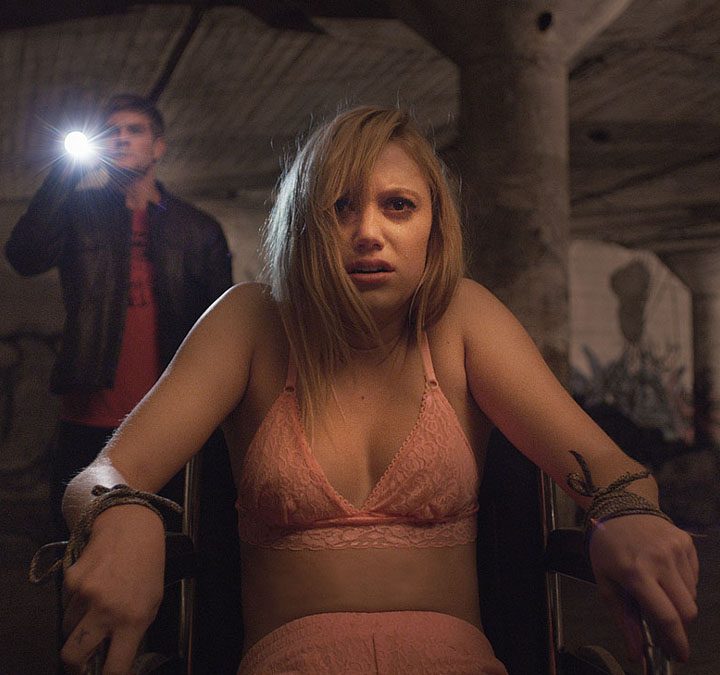 Maika Monroe bound to a chair with a man shining a flashlight behind her in It Follows.