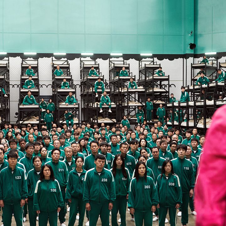 A worker in Squid Game standing in front of the many participants in numbered jumpsuits.