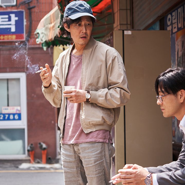 Seong Gi-Hun smoking a cigarette, Cho Sang-Woo sitting in a suit beside him from Squid Game.