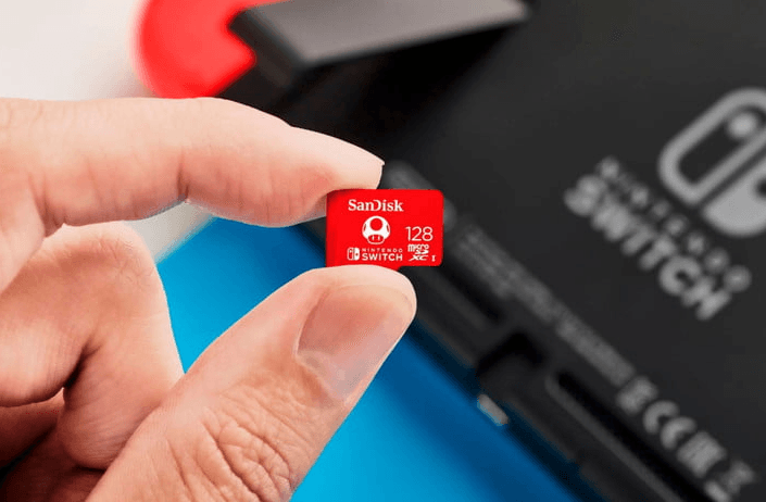 A hand holds a SanDisk 128GB microSDXC memory card for Nintendo Switch up close with console in background.