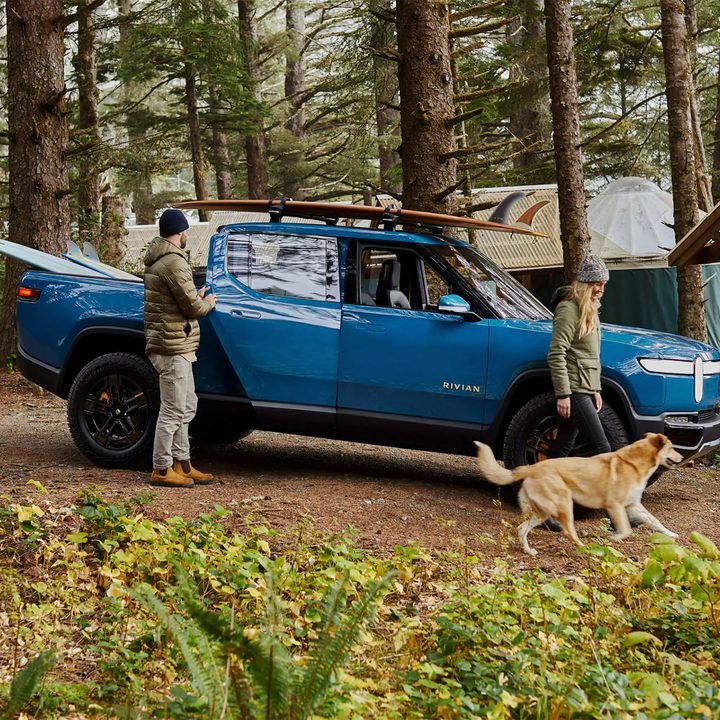 Blue Rivian R1T with a suftboard mounted on top while woman, man, and dog walk around it.