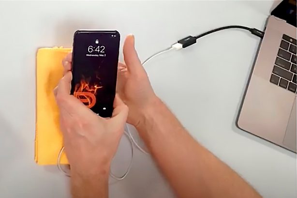 an iPhone held in the hand, connected to a Macbook via an adapter.