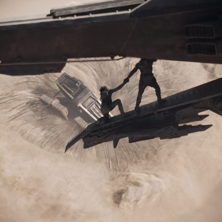 Sharacters from Dune look at the sand as a worm swallows a vehicle.