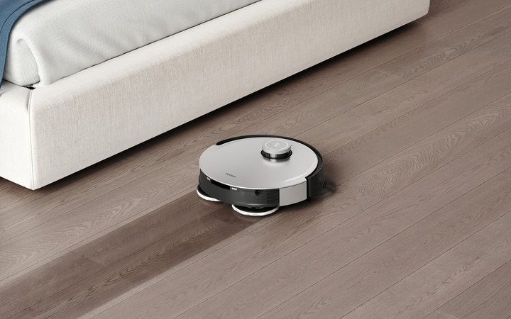 The Ecovacs Deebot X1 Omni cleans a hardwood floor in front of a bed.