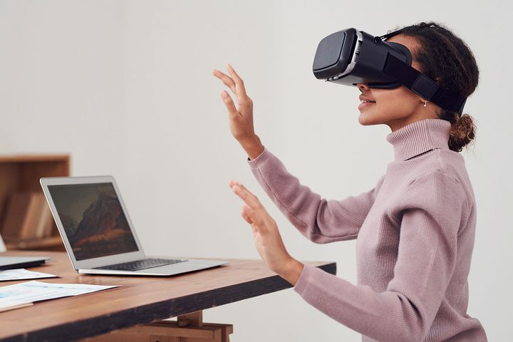 A woman using a VR headset at a desk with an Apple MacBook,
