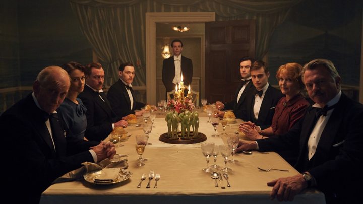 Guests seated at a dining table look at the camera in And Then There Were None.