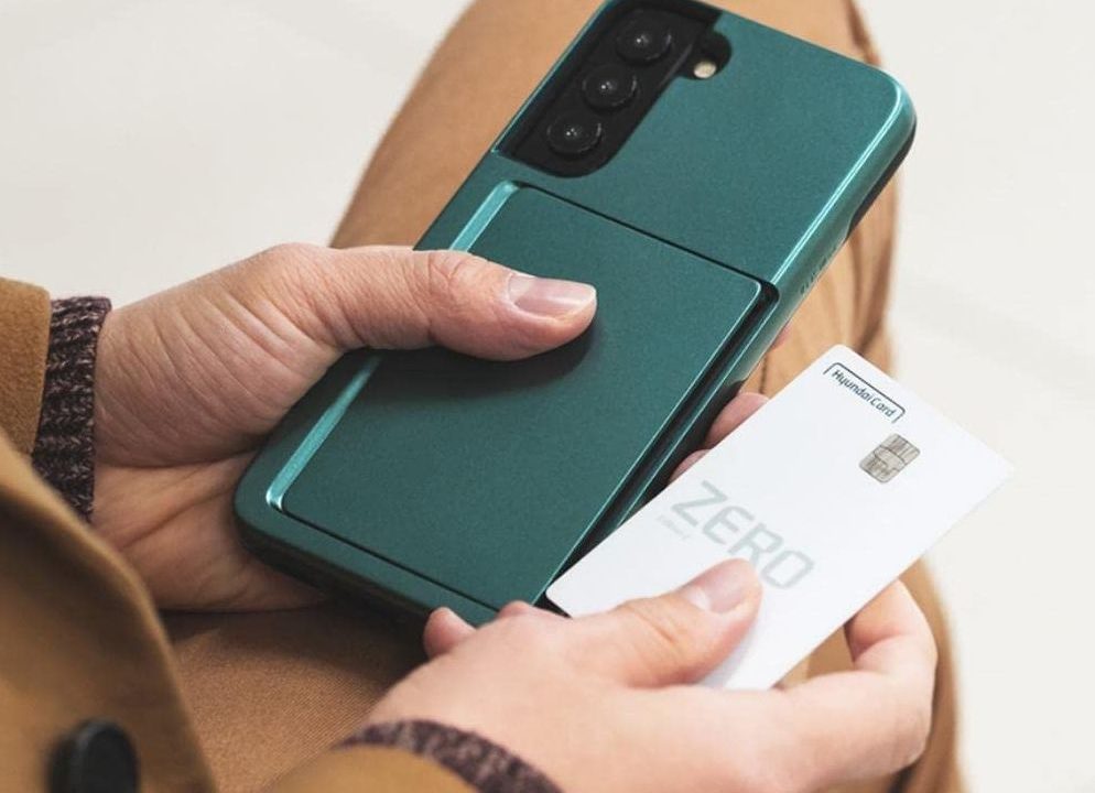 A person holding a card in one hand and the Galaxy S22 in a K-max auto-tok dark green card case in the other.