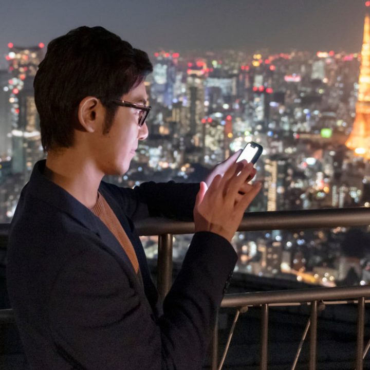 Man using smartphone while standing on balcony overlooking nighttime city skyline.
