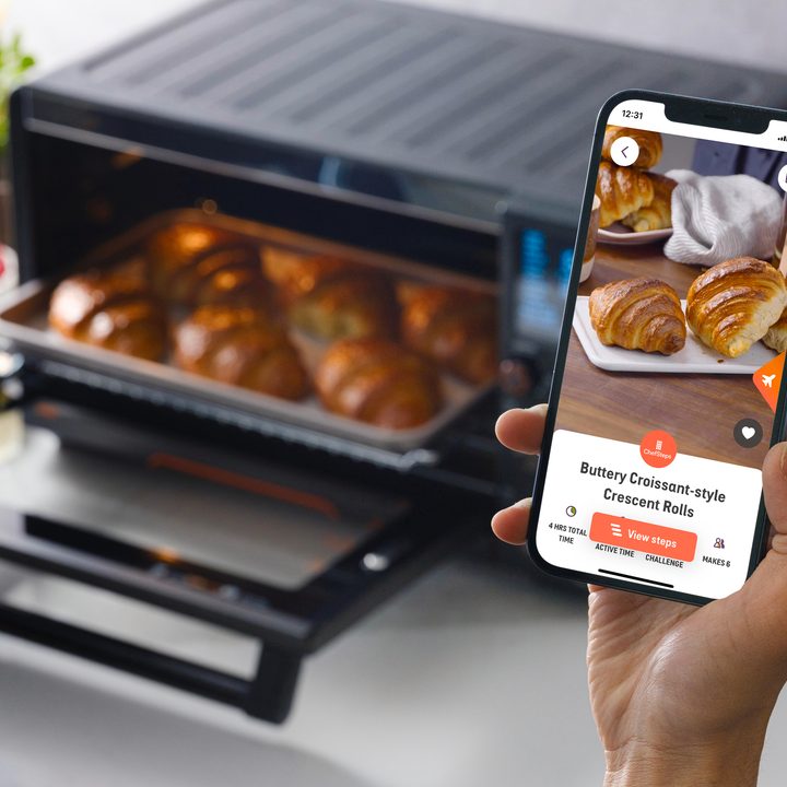 A hand holds a smartphone displaying the Joule Oven app with the Joule Oven full of croissants in the background.