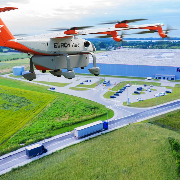 A rendering of the latest version of Elroy Air's autonomous aircraft flying over a roadway, with a building in the distance.