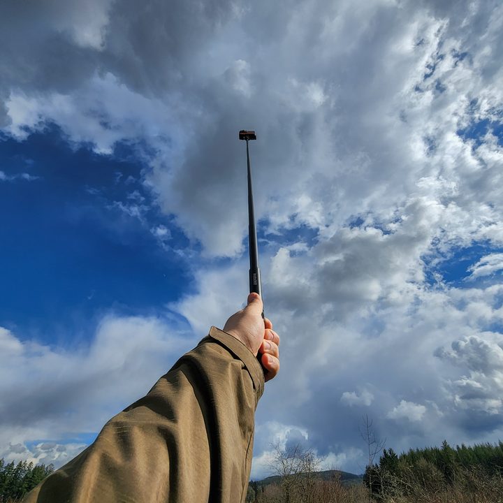 Insta360 One RS on selfie stick raised high into the air with a blue sky and clouds in the background.