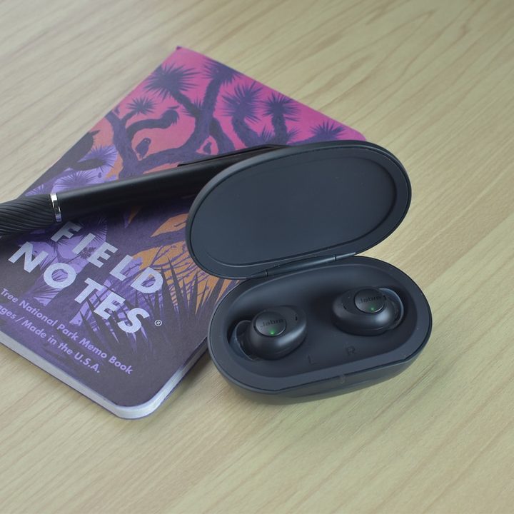 Jabra Enhance Plus earbuds in their open case, sitting next to a pen and a notebook.