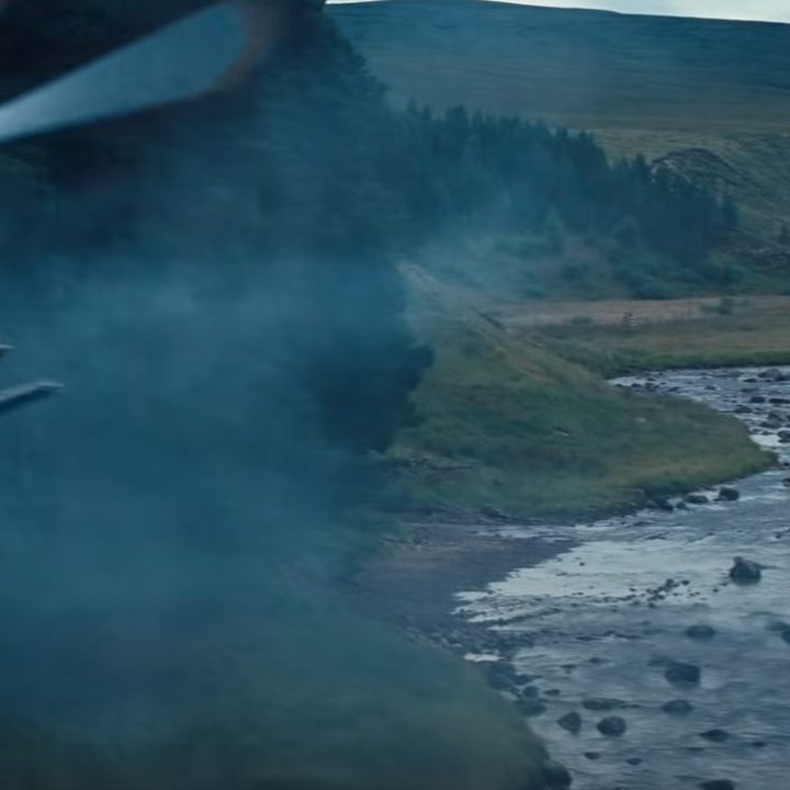 A car drives across a stream while a helicopter hovers nearby in a scene from No Time To Die.
