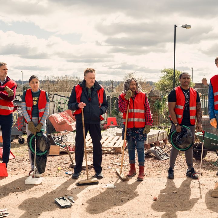 The cast of The Outlaws stands in a line with their community service vests on.