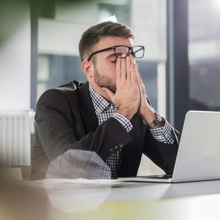 A tired-looking man sits at a laptop in an office.