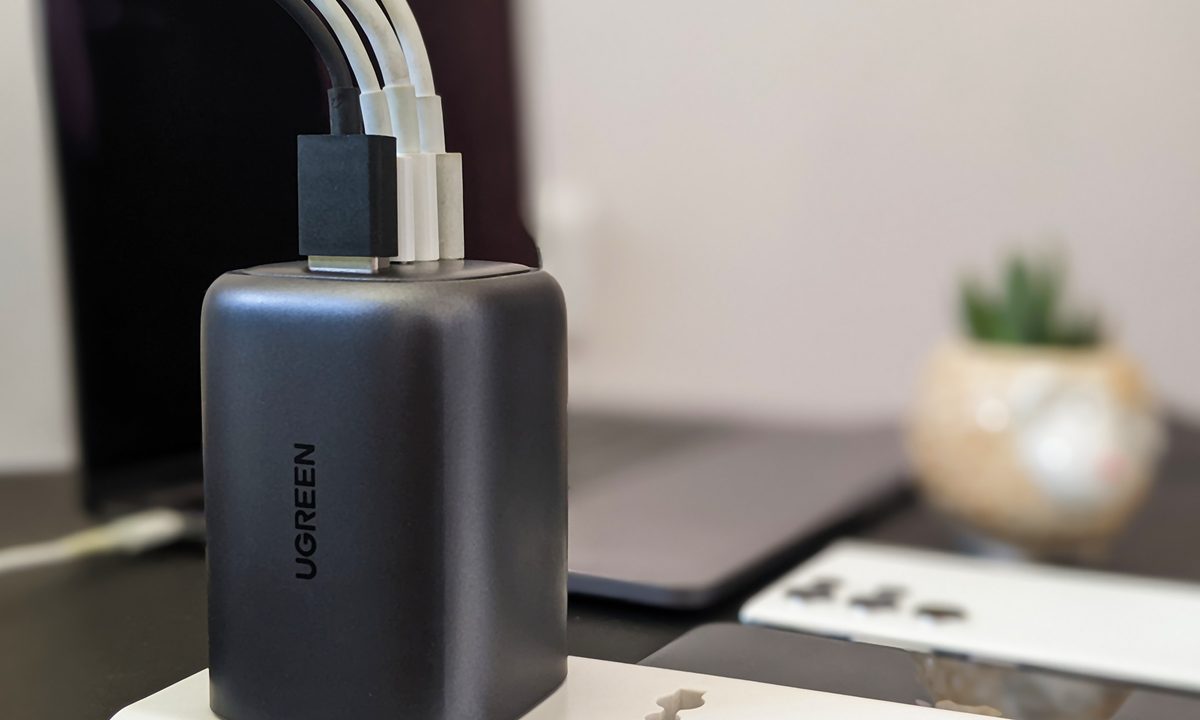 Ugreen 100 watts GaN gallium nitride charger with space gray Macbook, black iPhone 11, white Samsung Galaxy S22 Ultra, and a succulent in a pot on a black background.