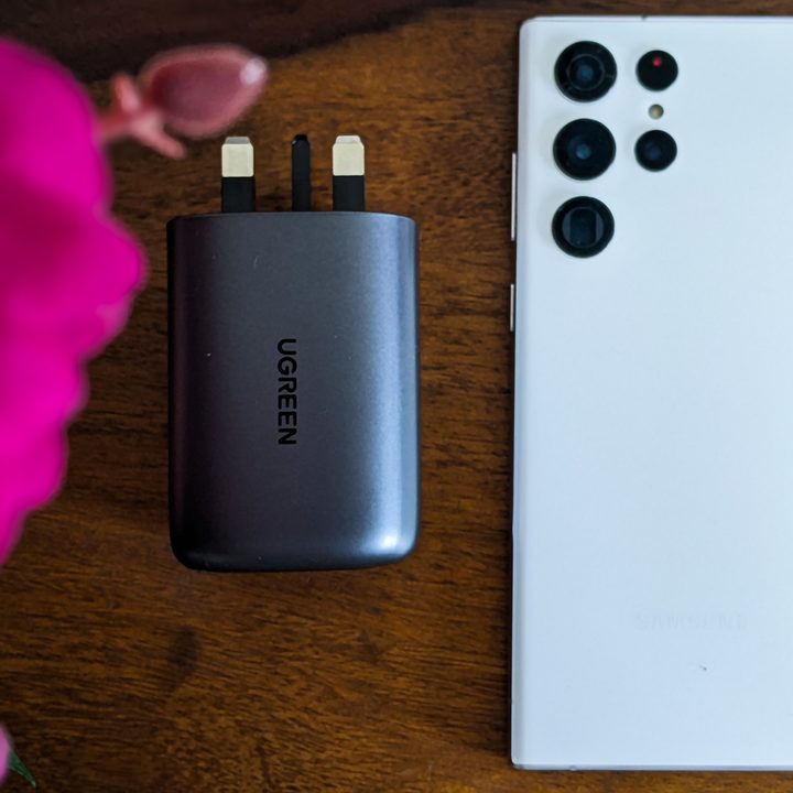The Ugreen 100W GaN gallium nitride fast charger next to a white Samsung Galaxy S22 Ultra on a table with pink flowers in the foreground.