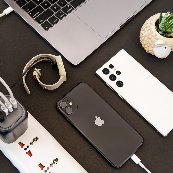 The Ugreen 100-watt GaN charger next to a space gray Macbook, black iPhone 11, white Samsung Galaxy S22 Ultra, and a succulent in a pot on a black background.