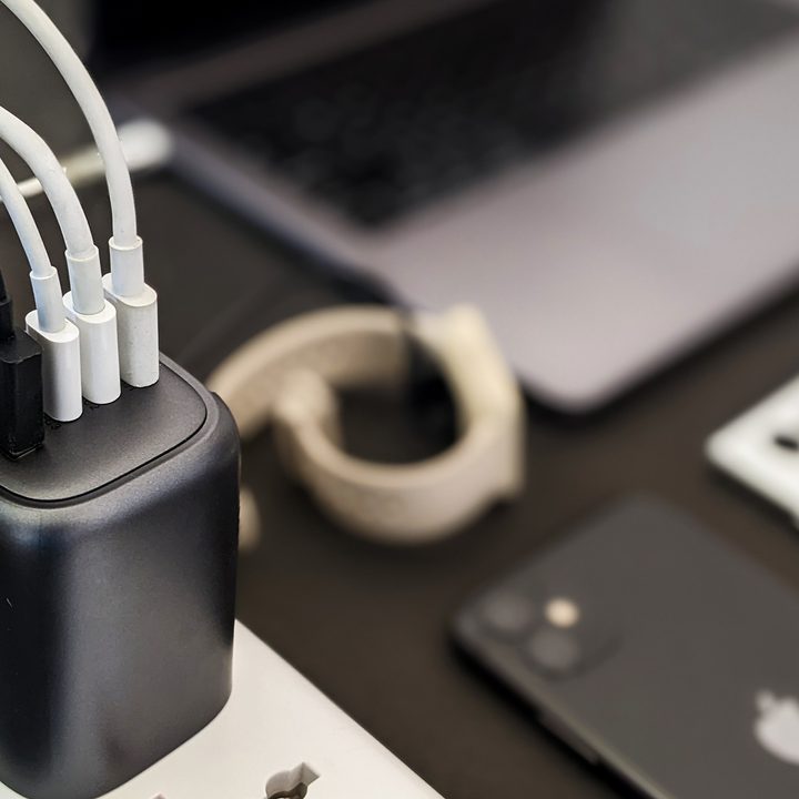 The Ugreen 100-watt GaN charger with a space gray Macbook, black iPhone 11, and white Samsung Galaxy S22 Ultra on a black background.