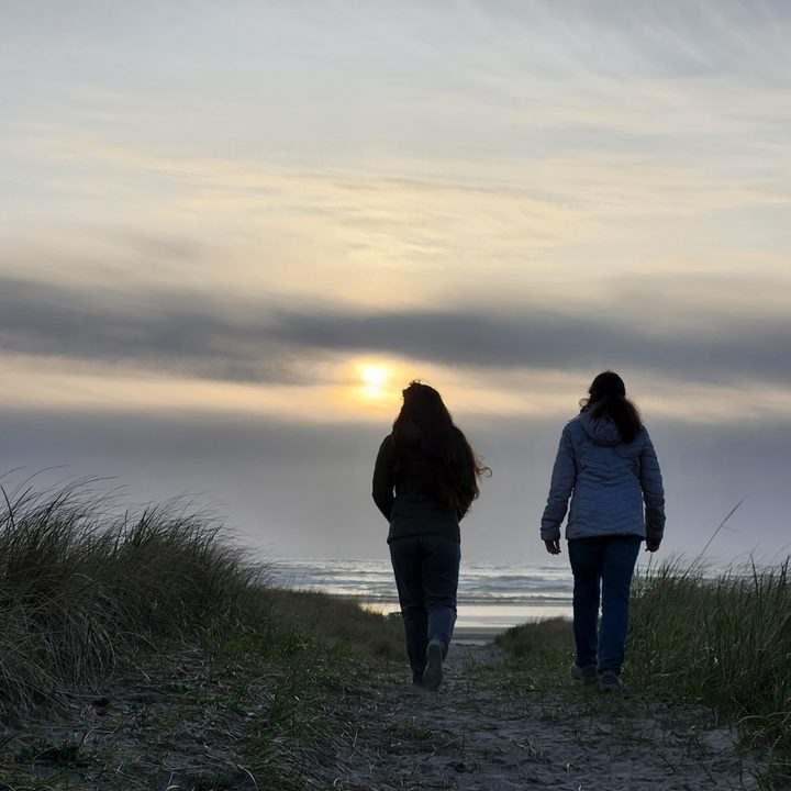 Two people walking over the top of a dune with the ocean and sunset in the background.