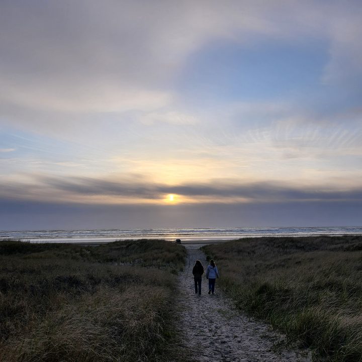 Sunset over dunes with grassy fields and two people walking on a path towards the ocean.