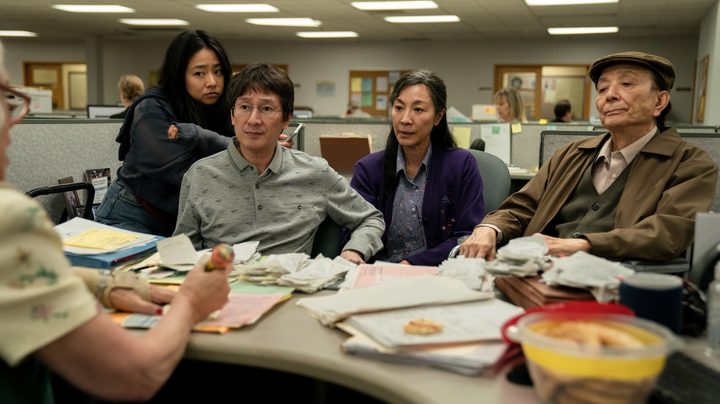 The cast of Everything Everywhere All At Once sits at a desk in an office building.