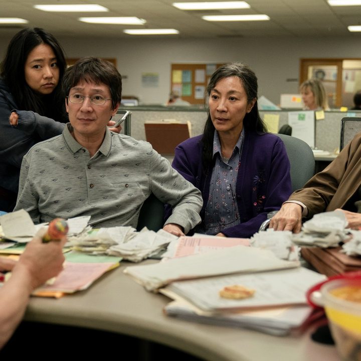 The cast of Everything Everywhere All At Once sits at a desk in an office building.