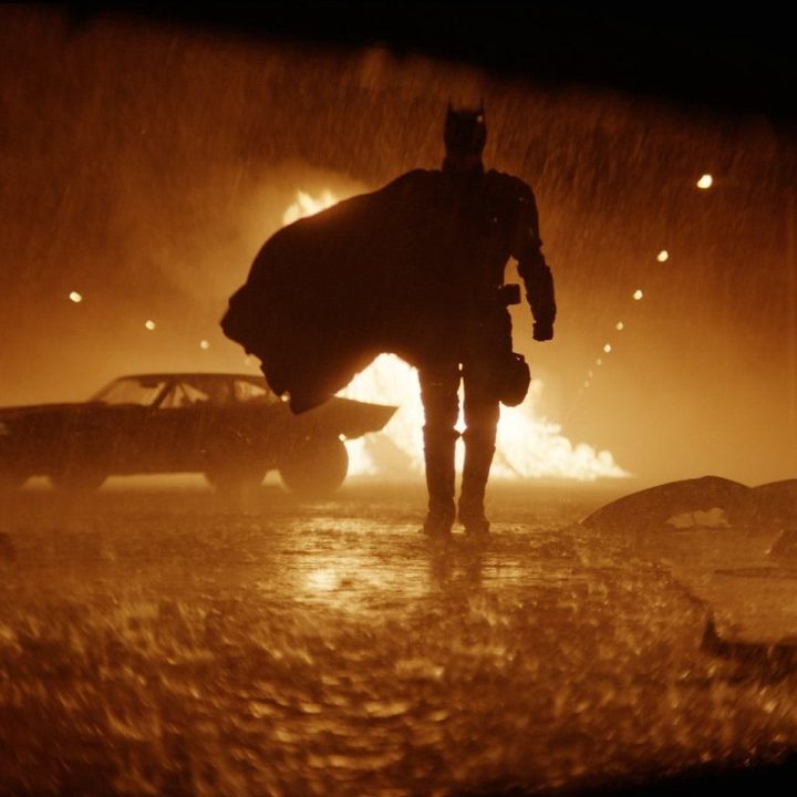 Batman approaches the camera on a rainy street with a fire in the background.